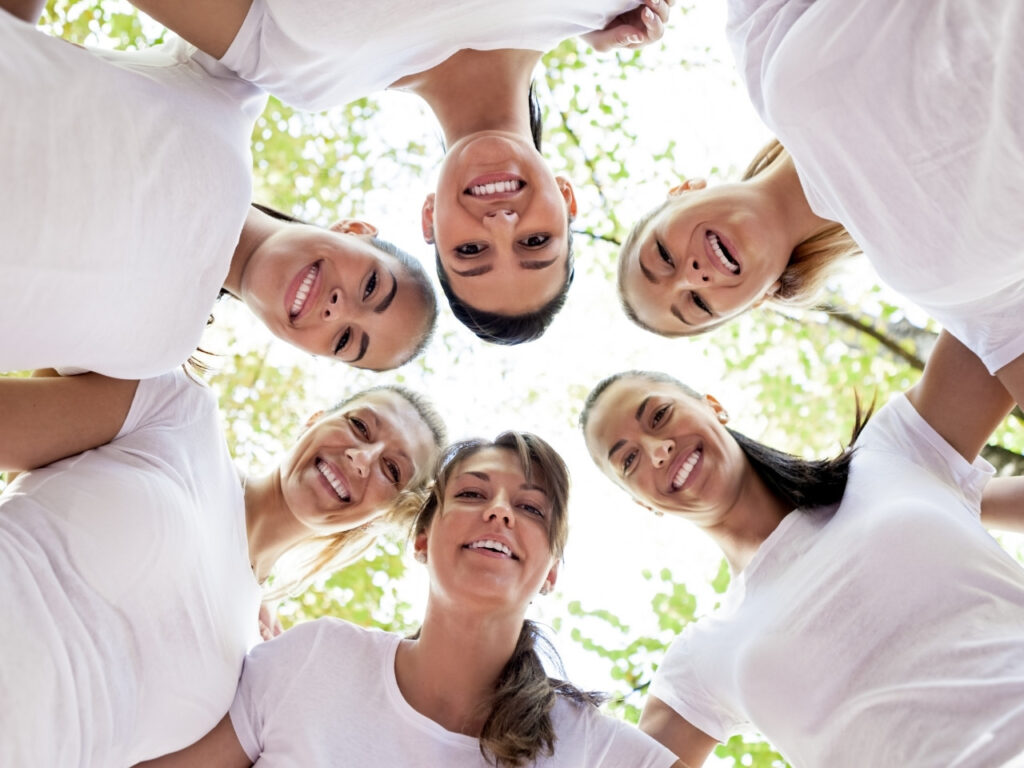 A group of smiling women standing in a circle, looking down at the camera with trees above them—symbolising connection, community and support during menopause.