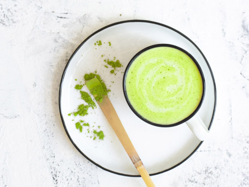 A creamy collagen matcha latte with a bamboo spoon and vibrant green matcha powder on a white plate – a nourishing midlife drink from The Menopause Kitchen.